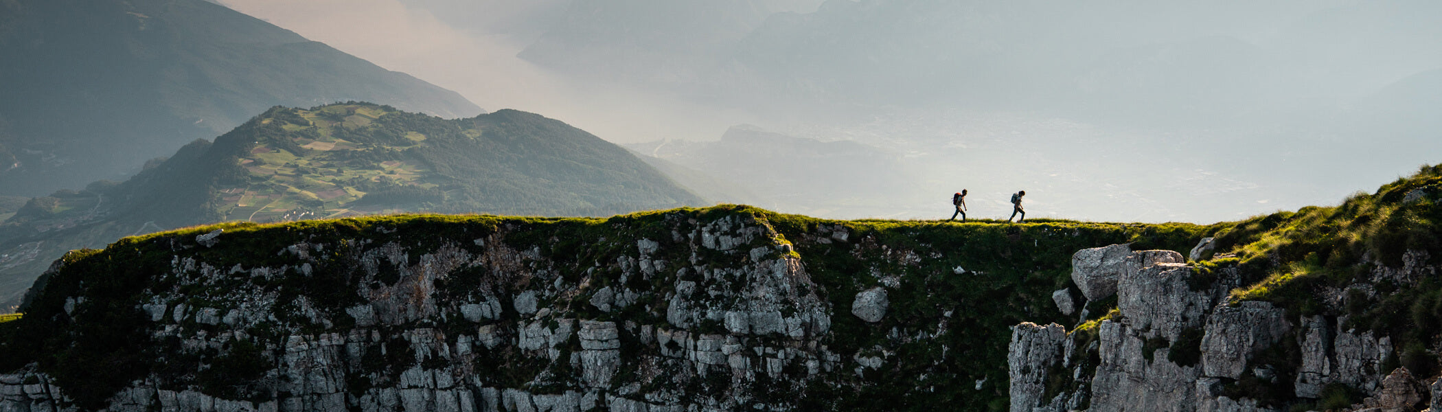 Two backpackers walking along a rocky cliff with misty mountains in the background. 
