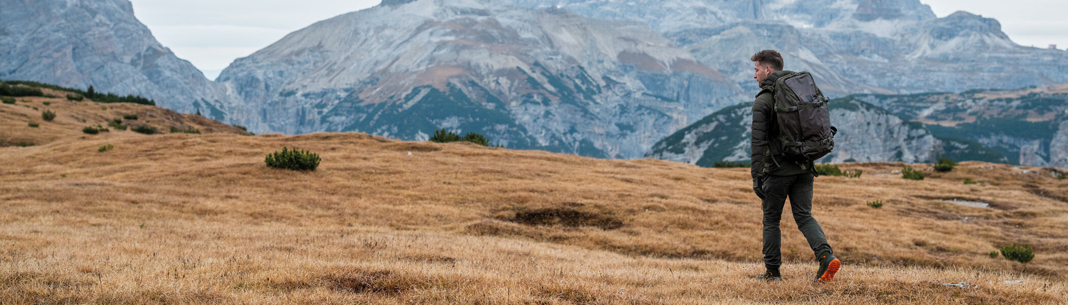 A man wearing outdoor gear walking in a field toward mountains. 