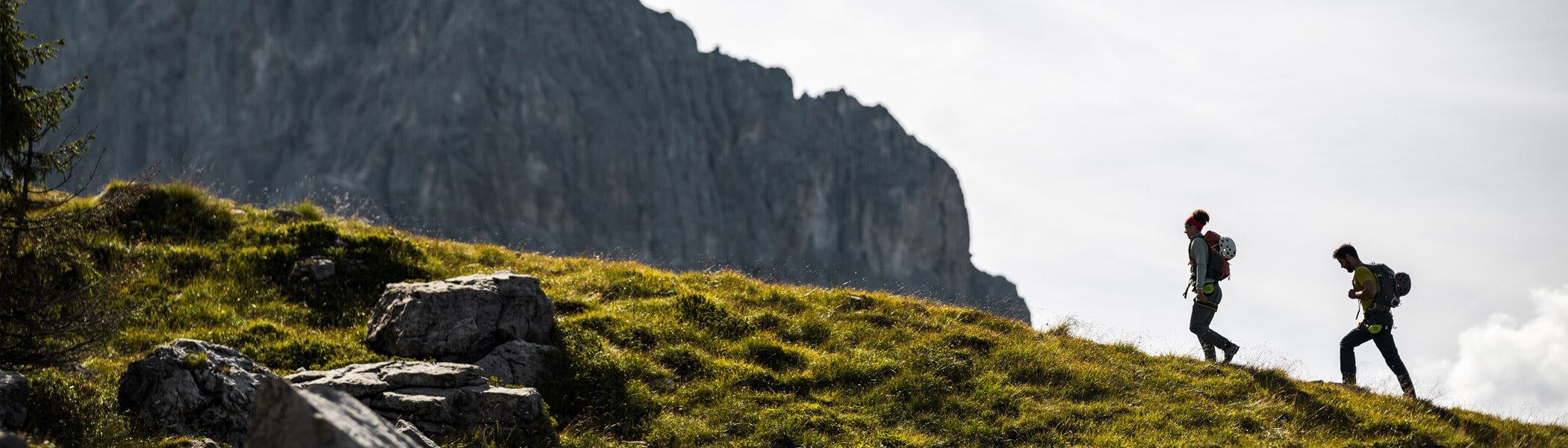 Two people hiking up a grassy ridge with the Italian Dolomite mountains in the background. 